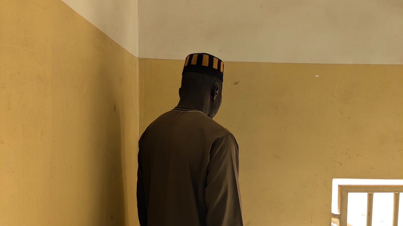 Man in a brown outfit and striped hat faces a beige wall, standing next to a window with bars letting in light.