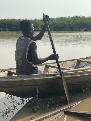 Person paddling a wooden boat on a calm river. Lush greenery in the background under a clear sky.