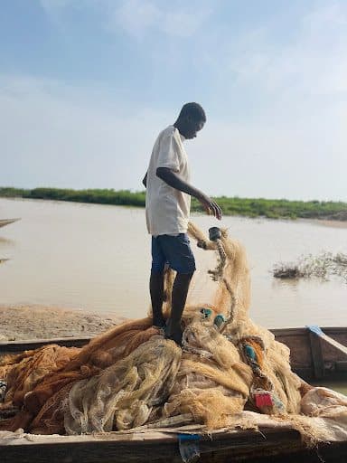 A person stands on a small boat, sorting fishing nets by a calm river or lake under a blue sky.